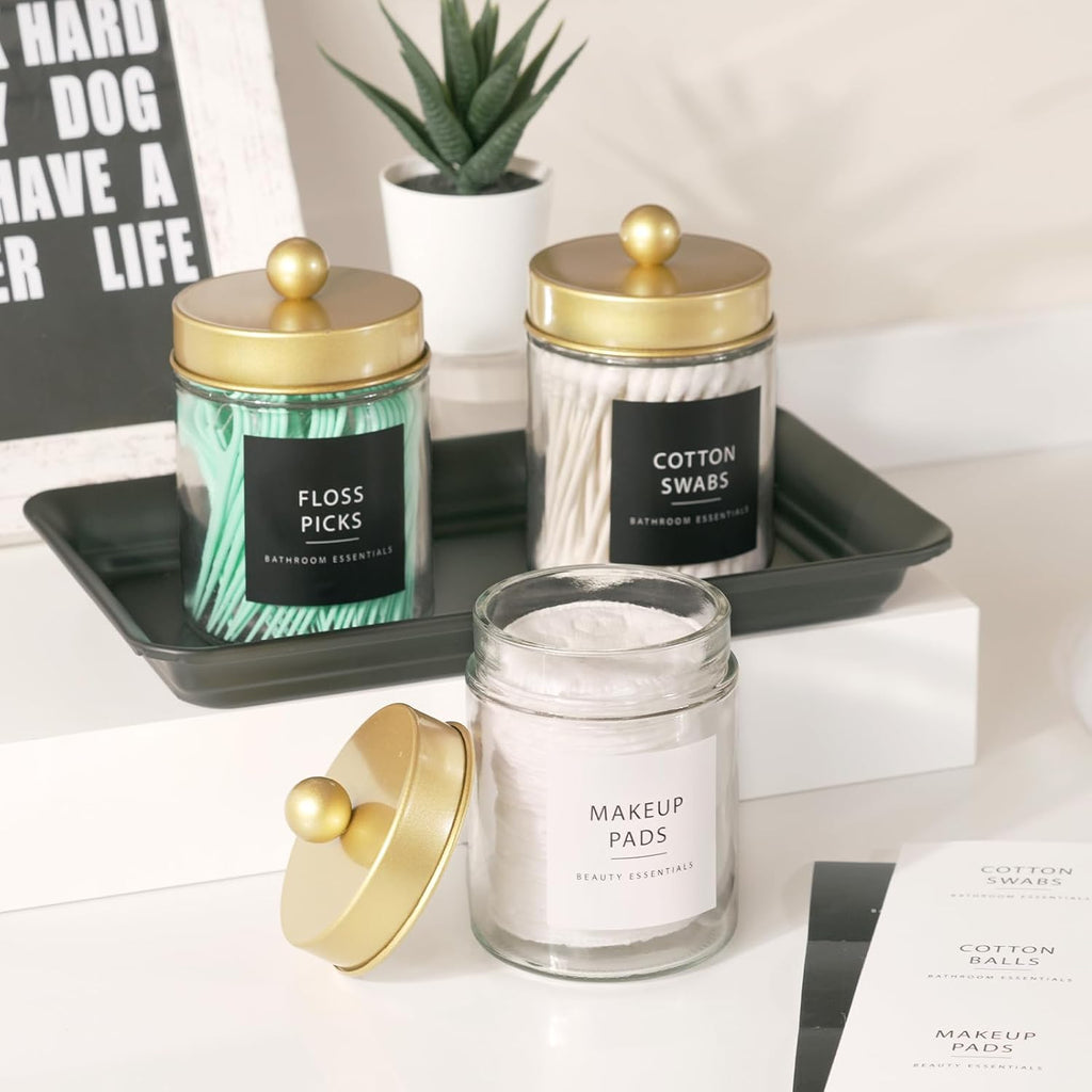 Gold-lid apothecary jars with floss picks, cotton swabs and makeup pads on a bathroom shelf