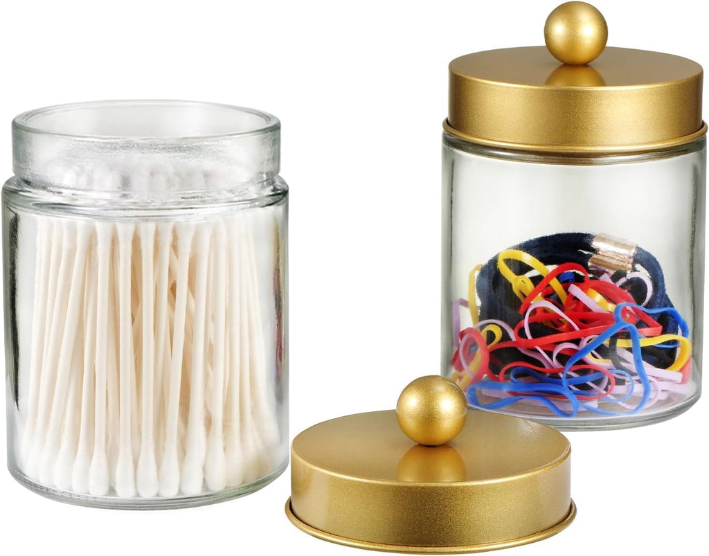 Glass apothecary jars holding cotton swabs and hair ties, one gold lid set aside