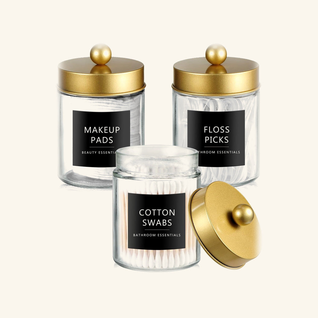 Three glass apothecary jars with gold lids holding makeup pads, floss picks and cotton swabs